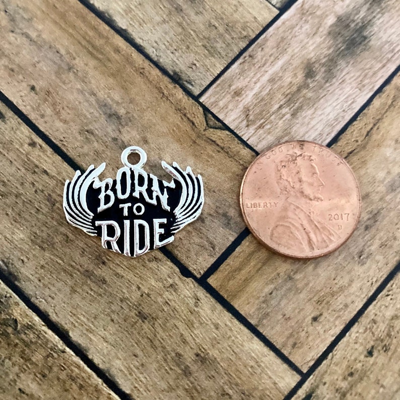 Puede incluir: Encanto de plata con alas negras y las palabras "Born to Ride" en letras blancas. El encanto es aproximadamente del tama&ntilde;o de una moneda de un centavo de d&oacute;lar estadounidense.