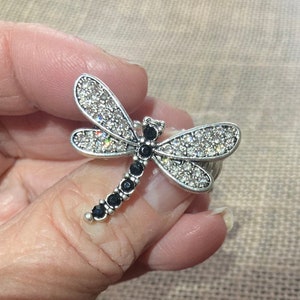 May include: A silver dragonfly ring with clear and black rhinestones. The dragonfly's wings are spread out and its body is adorned with black rhinestones.