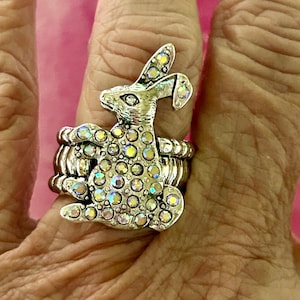 May include: A silver ring with a crystal-encrusted rabbit design. The rabbit is sitting up with its ears perked up. The ring is on a finger.