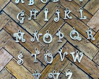 May include: A collection of twenty-six silver-toned metal alphabet letter charms, each featuring a decorative scrollwork design and a small loop at the top for attachment. The letters A through Z are arranged in rows, showcasing individual character styles.