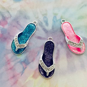 May include: Three silver charm pendants shaped like flip flops. The flip flops are decorated with glitter and rhinestones. The flip flops are blue, pink, and purple.