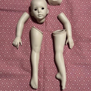 May include: A disassembled porcelain doll with a head, torso, two arms, and two legs. The doll parts are off-white and arranged on a pink fabric with a white star pattern. The doll's face has black eye sockets.