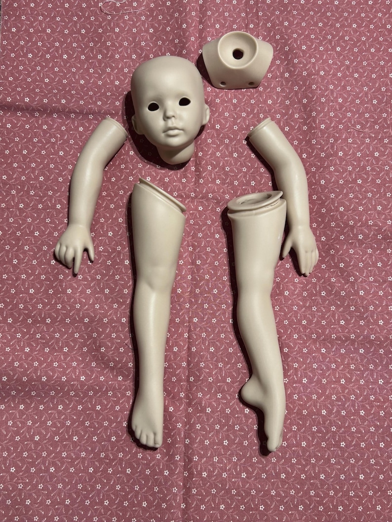 May include: A disassembled porcelain doll with a head, torso, two arms, and two legs. The doll parts are off-white and arranged on a pink fabric with a white star pattern. The doll's face has black eye sockets.
