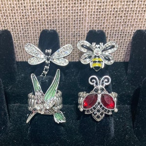 May include: Five silver rings with different animal designs. The rings feature a dragonfly, a bee, a hummingbird, and two ladybugs. The rings are all set with rhinestones and have a silver finish.