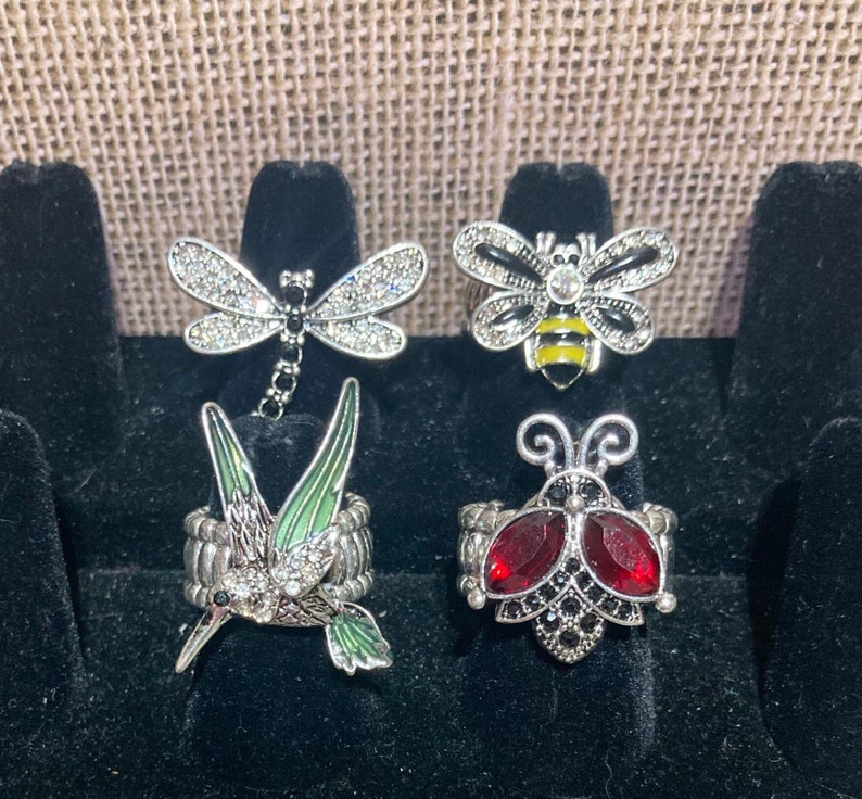 May include: Five silver rings with different animal designs. The rings feature a dragonfly, a bee, a hummingbird, and two ladybugs. The rings are all set with rhinestones and have a silver finish.