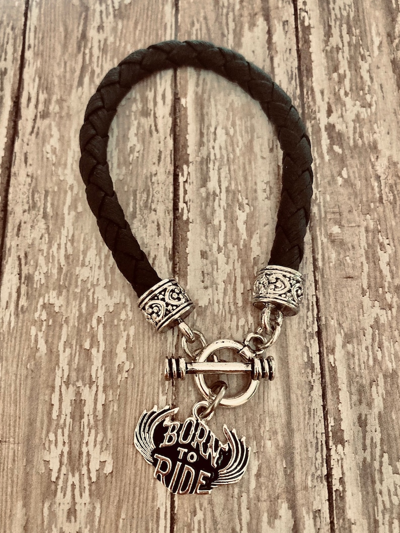 Puede incluir: Pulsera trenzada de cuero negro con cierre de palanca plateado y un dije plateado que dice "Born to Ride" con alas.