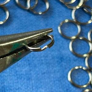 May include: Close-up of silver jump rings being closed with pliers on a blue surface.