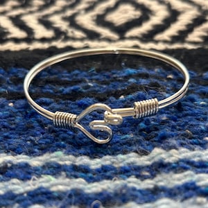 May include: A silver-colored bangle bracelet with a heart-shaped clasp. The bracelet features a wire-wrapped design on both sides of the clasp. The background is a blue and black patterned fabric.
