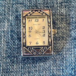 May include: A rectangular silver-toned quartz watch with the text "Abby Girl" on the face. The watch has a cream-colored face with black numerals and hands. The watch is set against a blue denim background.