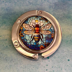 May include: A circular silver-toned metal bag hanger featuring a central glass cabochon insert. The insert displays a detailed illustration of a bee centered against a blue and yellow stained glass-style honeycomb pattern with small floral accents.