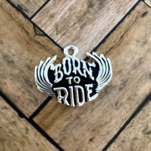 Puede incluir: Encanto de plata con alas negras y las palabras "Born to Ride" en una fuente estilizada.