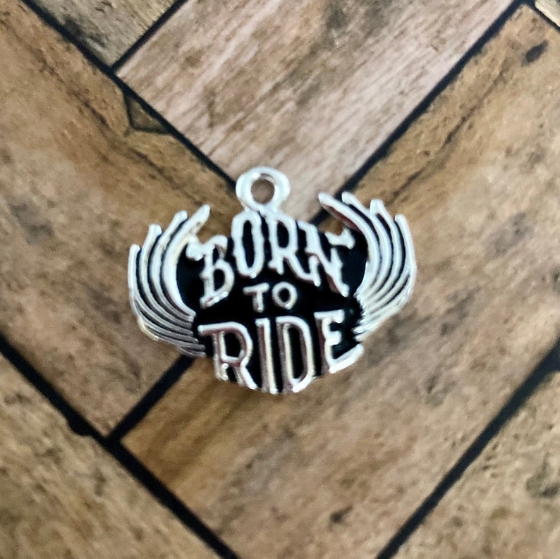Puede incluir: Encanto de plata con alas negras y las palabras "Born to Ride" en una fuente estilizada.