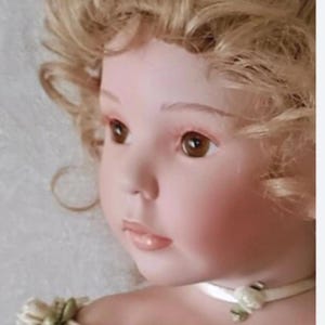 May include: A porcelain doll with a fair complexion, rosy cheeks, and brown eyes. The doll has curly blonde hair and is wearing a white ribbon choker with a floral accent. The doll's face is detailed with painted features.