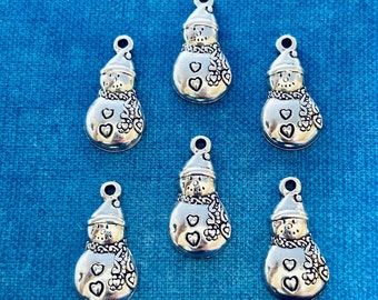 May include: Six silver-colored snowman charms with a loop at the top for hanging. Each snowman has a hat, scarf, and heart details. The charms are displayed on a blue background.