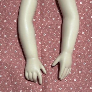 May include: Two porcelain doll arms with hands, in a cream color. The arms are slightly bent at the elbow and the hands are open. The arms are on a pink fabric with a white floral pattern.