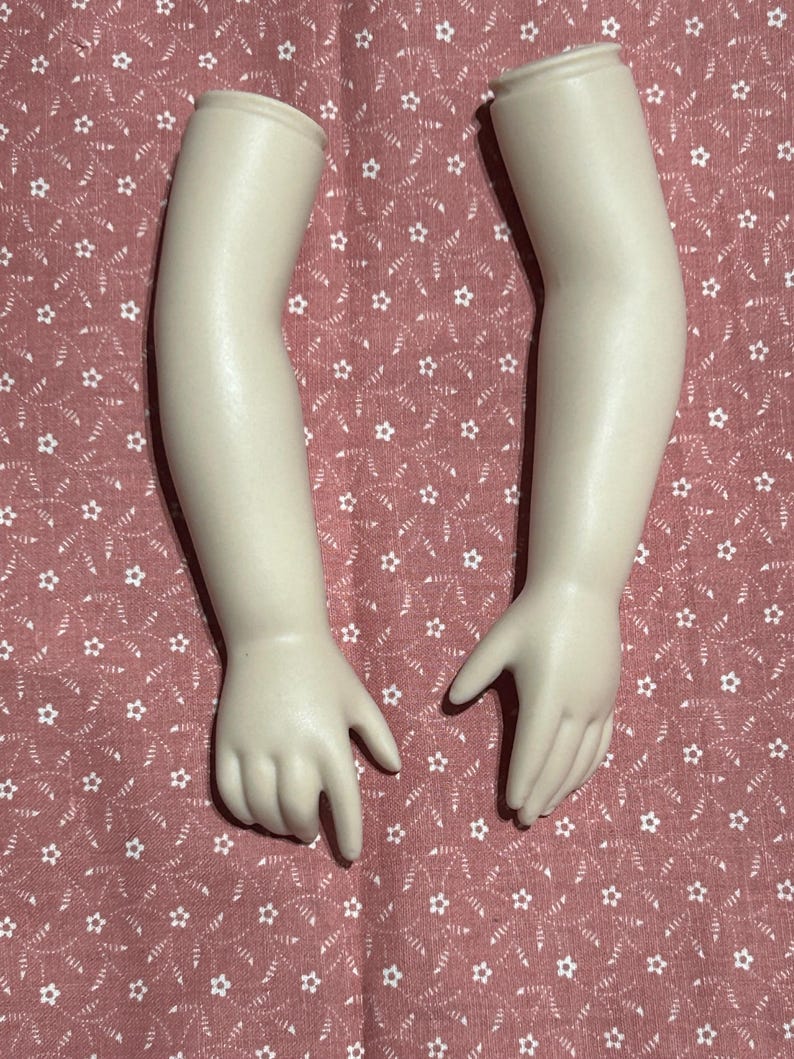 May include: Two porcelain doll arms with hands, in a cream color. The arms are slightly bent at the elbow and the hands are open. The arms are on a pink fabric with a white floral pattern.