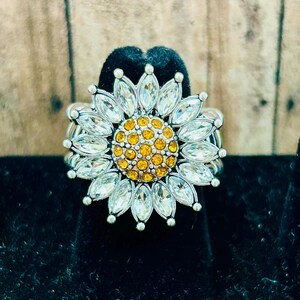 May include: A silver ring with a white rhinestone sunflower design. The center of the sunflower is filled with yellow rhinestones.