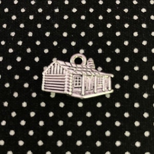 May include: A silver charm in the shape of a log cabin with a small chimney and windows. The charm has a loop at the top for attaching to a bracelet or necklace.
