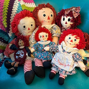 May include: Five Raggedy Ann dolls, each with a different dress and hair style. The dolls are all red-haired and have a sweet, innocent expression. The dolls are sitting on a blue surface.