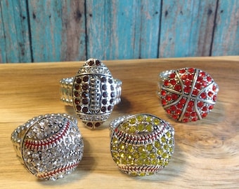 May include: Four adjustable rings featuring different sports ball designs. The rings are silver with rhinestones and feature a baseball, basketball, football, and softball design.