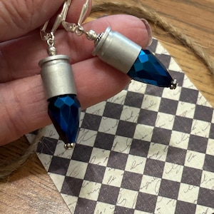 May include: A pair of silver-toned leverback earrings. Each earring features a silver-colored cylindrical top and a faceted, deep blue, teardrop-shaped crystal. The earrings are displayed against a checkered background.