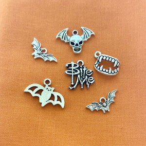 May include: A set of six silver charm pendants featuring Halloween themes. The charms include a bat, a skull, a vampire fang, and the words "Bite Me".