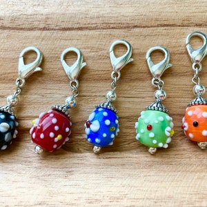 May include: Five colorful glass beads with white polka dots and silver metal clasps. The beads are attached to silver chains and are used as stitch markers for knitting or crocheting.