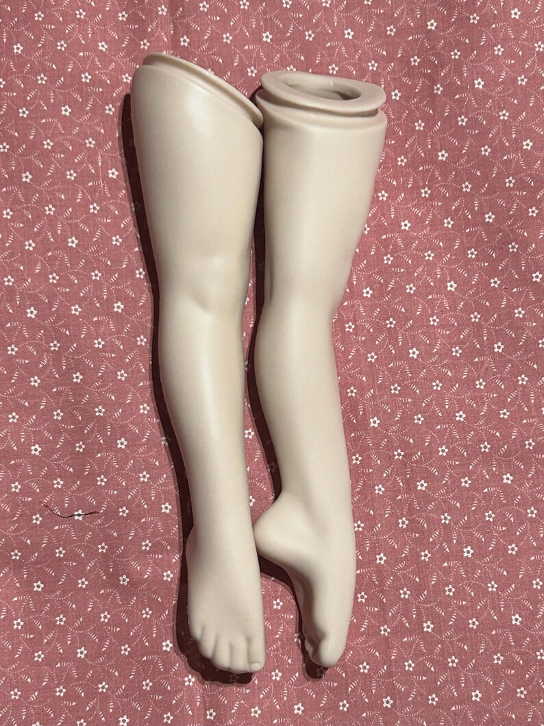 May include: Two porcelain doll legs, off-white in color, are displayed against a red background with a white star pattern. The legs are smooth and detailed, with visible toes and a slight bend at the knees. The top of each leg has a cylindrical opening.