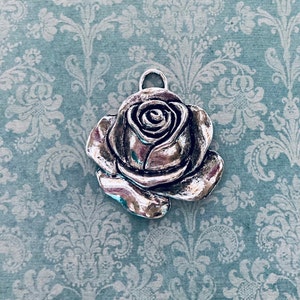 May include: A silver-colored rose pendant with a loop for hanging. The rose has detailed petals and a textured surface. The pendant is set against a light blue background with a floral pattern.