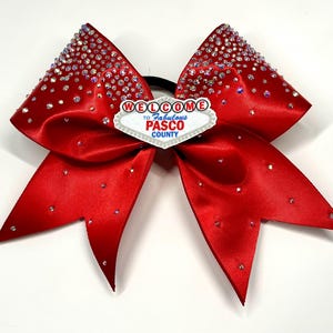May include: A red cheerleading bow with sparkling rhinestones and a black elastic band. The bow features a white and silver emblem that reads "Welcome to Fabulous Pasco County."