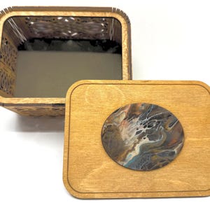 May include: A rectangular wooden box with an open top and a separate lid. The box features laser-cut decorative side panels. The lid is made of light brown wood and features a circular inlay with a marbled, multi-colored paint pour design.