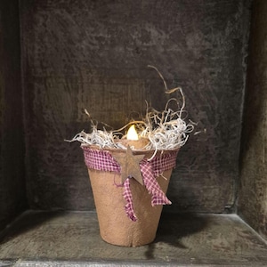 May include: A rustic candle holder with a warm, glowing LED candle. The holder is a brown pot, adorned with a burgundy and white plaid ribbon and a star-shaped embellishment. The top is filled with decorative straw.