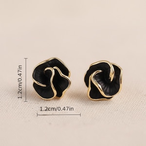 Könnte beinhalten: Ein Paar schwarze Ohrstecker in Blumenform mit goldfarbenem Rand. Jeder Ohrring misst 1,2 cm im Durchmesser. Die Ohrringe sind vor einem neutralen Hintergrund platziert.