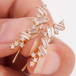 Puede incluir: Un par de pendientes trepadores dorados con piedras de circonita c&uacute;bica transparentes, de corte rectangular y redondo. Los pendientes tienen un dise&ntilde;o delicado y moderno.