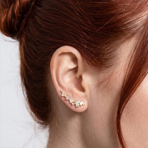 Puede incluir: Un pendiente trepador dorado con m&uacute;ltiples piedras transparentes, de corte rectangular y redondo. El pendiente se lleva en la oreja de una persona con cabello casta&ntilde;o rojizo.