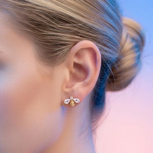 May include: A gold-coloured bee stud earring with clear, sparkling accents. The bee's wings are adorned with clear crystals, and the body has a textured, honeycomb design. The earring is worn on an ear, with a soft, blurred background.