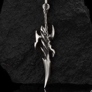 May include: A silver pendant in the shape of a stylized sword, suspended from a black cord. The pendant has intricate, sharp details and a polished finish. The background is a dark, textured surface. The pendant is approximately 3 inches long.