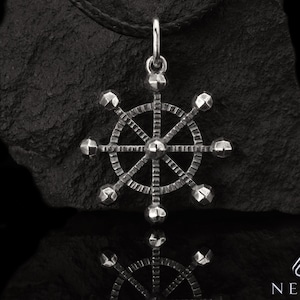 Könnte beinhalten: Ein silberfarbener Anhänger in Form eines Schiffsrads, der an einer schwarzen Kordel hängt. Das Raddesign weist Speichen und abgerundete Knöpfe auf. Der Anhänger wird vor einem dunklen, strukturierten Hintergrund präsentiert.