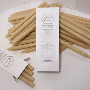 Puede incluir: Velas para oídos de cera de abeja beige, algunas en una caja blanca abierta, otras dispersas. Una tarjeta blanca muestra "Nature's Beeline" e información del producto. Las velas cilíndricas son de materiales naturales.