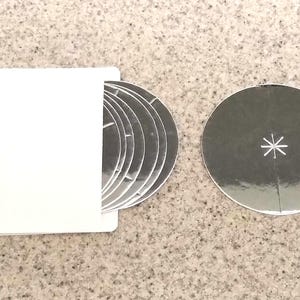 Puede incluir: Un juego de posavasos circulares plateados con una funda de papel blanca. Un posavasos está separado, mostrando un diseño de estrella. Los posavasos son de material reflectante, y la funda es blanca.