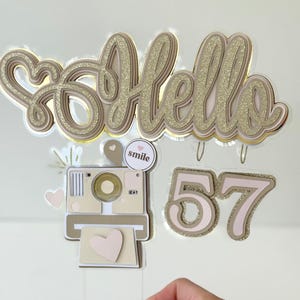May include: A layered cake topper with the word "Hello" in gold glitter script, a camera, hearts, and the number "57". The camera and numbers are in shades of tan and pink. The topper is designed for a birthday celebration.