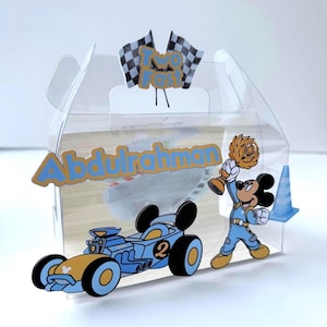 Puede incluir: Una caja de regalo de plástico transparente decorada con un tema de carreras. La caja presenta un coche de carreras azul, un personaje de dibujos animados sosteniendo un trofeo y el texto "Two Fast" y "Abdulrahman". La caja está decorada con banderas a cuadros y un cono de tráfico.