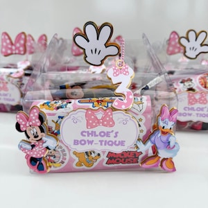 Puede incluir: Cajas transparentes para recuerdos de fiesta decoradas con el tema de Minnie Mouse. Cada caja tiene una etiqueta rosa con el texto "Chloe's Bow-Tique" y personajes de dibujos animados. Un dije con el número 3 y recortes de manos de Mickey Mouse están adjuntos.