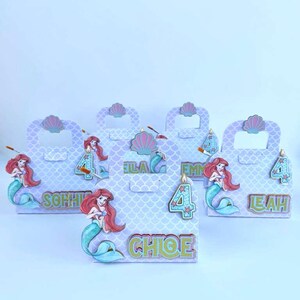 May include: A set of five light purple gift boxes with a mermaid scale pattern. Each box features a cutout handle, a pink seashell accent, a cartoon mermaid illustration, a number four, and a personalized name in green and gold lettering.