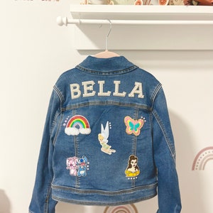 Puede incluir: Una chaqueta vaquera azul con el nombre "BELLA" bordado en la espalda. La chaqueta tiene varios parches coloridos, incluyendo un arcoíris, una mariposa, un hada y un personaje de dibujos animados.