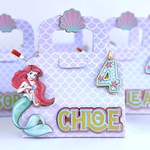 May include: A light purple paper gift bag with a scalloped scale pattern, featuring a cutout handle topped with a pink seashell. The bag displays a mermaid illustration, a blue number 4 candle, and the name CHLOE in gold glittery letters.