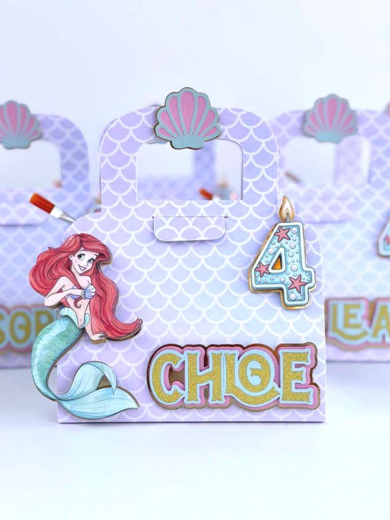 May include: A light purple paper gift bag with a scalloped scale pattern, featuring a cutout handle topped with a pink seashell. The bag displays a mermaid illustration, a blue number 4 candle, and the name CHLOE in gold glittery letters.