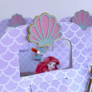 May include: A light purple paper gift bag featuring a repeating white scale pattern. The top center is decorated with a layered seashell cutout in pink and blue with a gold border. Through the cutout window, a cartoon character with red hair is visible.