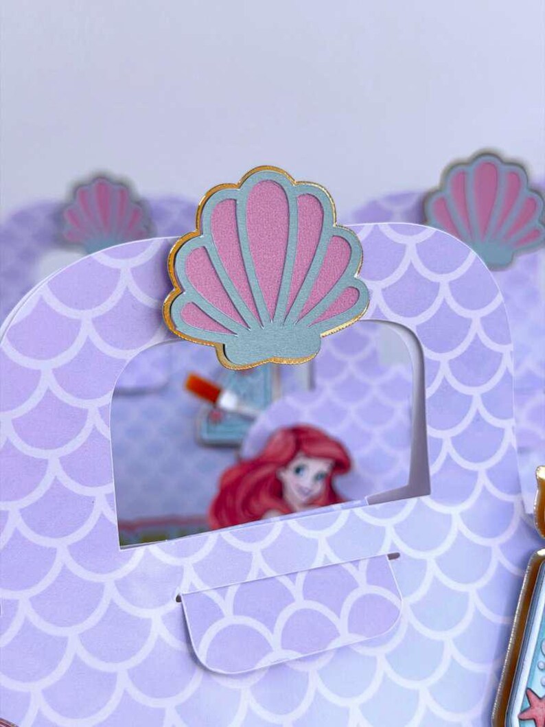 May include: A light purple paper gift bag featuring a repeating white scale pattern. The top center is decorated with a layered seashell cutout in pink and blue with a gold border. Through the cutout window, a cartoon character with red hair is visible.