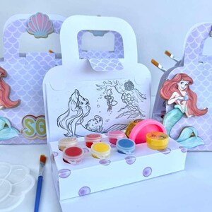 May include: A white cardboard craft box with a handle, featuring a mermaid illustration and a black and white coloring scene. The box contains small circular paint pots in red, yellow, blue, and pink, alongside a pink container of modeling dough.